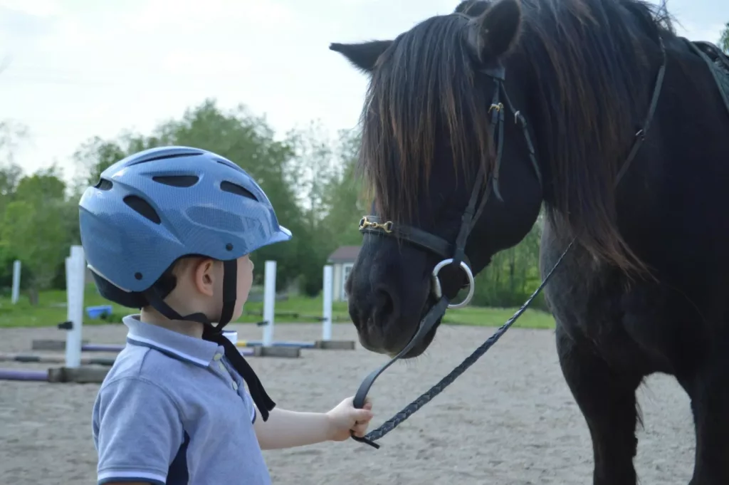 Un enfant casqué tient par les rênes un cheval noir dans une carrière